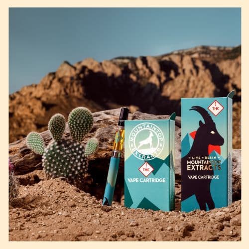 a photo of a cartridge on a battery and vape packaging by Mountaintop Extracts. the cartridge and boxes are sitting in a New Mexico desert with a cactus and the Sandi Mountains in the background.