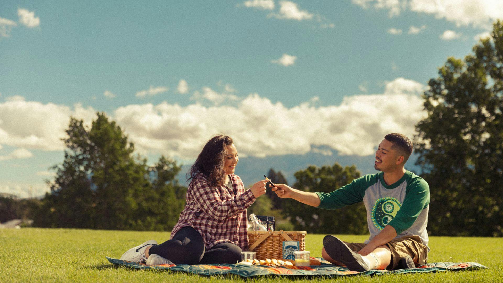 a photo of a woman and a man sitting on a picnic blanket in a field of green grass with blue skies and fluffy white clouds. he is handing her a cannabis vape pen and in addition to picnic snacks, there is a package of Oasis cannabis powder on the blanket.