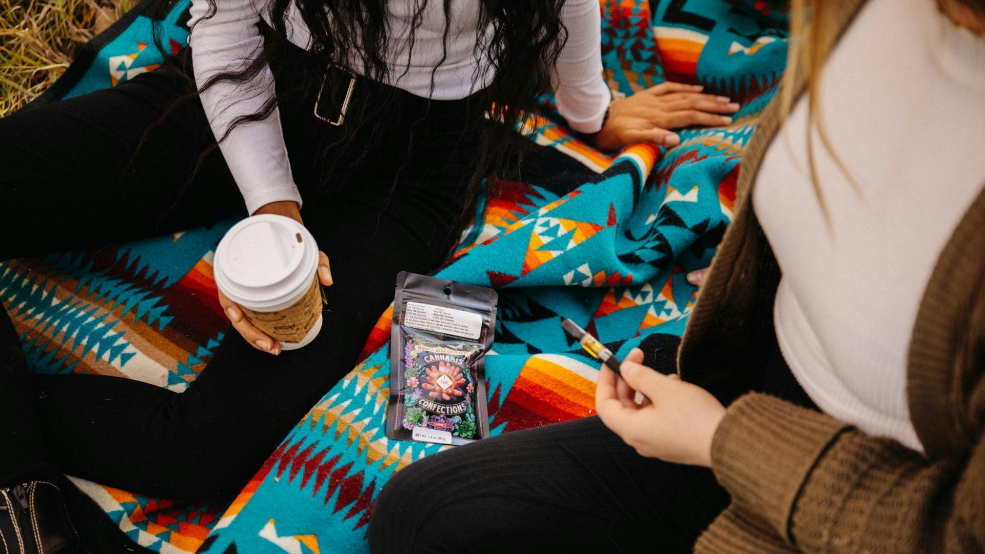 photo of two people having a picnic. you see them from the shoulders down, one is holding a cup of coffee, one is holding a cannabis vape  pen. there is a package of edibles from Mountaintop Extracts on the teal blanket.